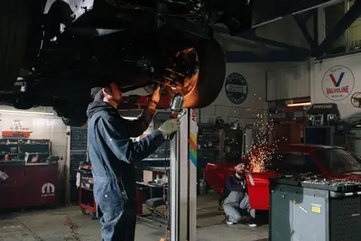 Two automotive technicians repairing a car in a busy workshop with sparks flying - Repair People You Need to Know, Just in Case