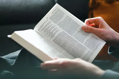 Close-up of a person's hands turning the pages of a Bible open to the Gospel of Matthew, seated on a dark sofa - 7 Proven Strategies To Become A More Insightful Reader Of Books
