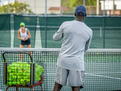Tennis coach with ball basket watching player during practice session - How to Become a Tennis Coach: A Step-By-Step Guide