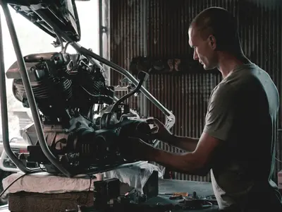 Man in a garage examines an exposed motorcycle engine and drivetrain mounted on a workbench, with a corrugated metal wall behind him - Bike Maintenance Tips for Every Rider