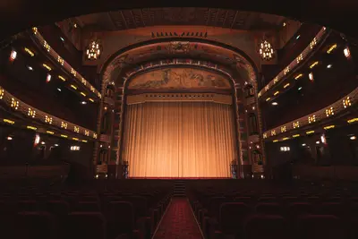 An ornate art deco theatre auditorium with a lit gold stage curtain, tiered balconies decorated with patterned lighting, and rows of empty red seats leading to the stage - From Local Stage to Global Spotlight