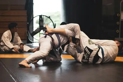 Two Brazilian jiu-jitsu practitioners in white gis grappling on the mat, one attempting an armbar from the top position, with other students seated and training in the background - How Self-Defence Skills Empower Individuals and Build Confidence