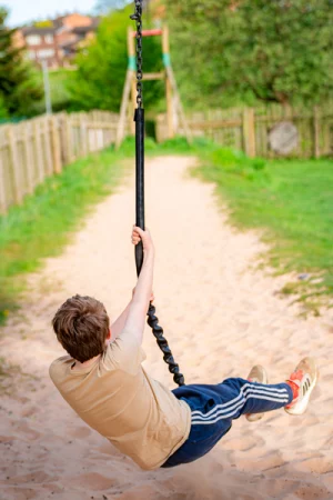 The Land, Caia Park, Wrexham - A boy rides a zip line along a sandy playground path, viewed from behind, with wooden fencing, grass and trees lining the route - Charity News: AVOW, Young Carers & Marie Curie