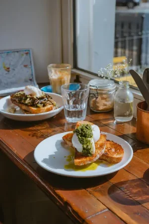 Burrata and pesto on toasted sourdough drizzled with olive oil, on a sunlit café table with a second dish of mushroom toast and poached egg, an iced coffee, water glasses and a small flower jar in the background - Why Brunch Became the Ultimate Weekend Ritual