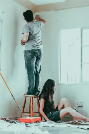 A man stands on a stool painting near the ceiling while a woman sits on the dust-sheeted floor below, in a room being redecorated with a paint tray, roller and stripped window frame - How Delaying Home Repairs and Maintenance Ends up Costing You