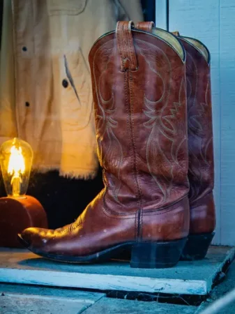A close-up of a pair of brown leather cowboy boots with stitched western detailing, lit warmly beside a vintage-style Edison bulb lamp - Country & Western Night Ebenezer Cefn Mawr