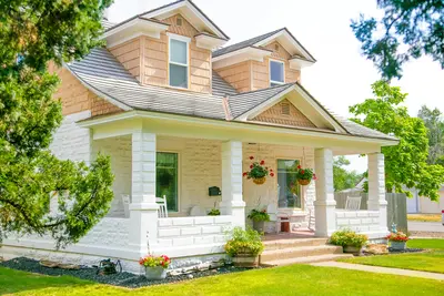 Cream stone bungalow with cedar-shingled gables and dormer windows, a white-columned wraparound porch with rocking chairs and hanging geranium baskets, framed by mature trees and a landscaped front lawn. - Boosting Curb Appeal: Simple Upgrades for Your Home