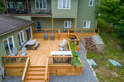 Aerial view of a large multi-level wood deck behind a green house, with a dining table, Adirondack chairs, a built-in BBQ, planter boxes, and a stone staircase leading down to a landscaped garden bed - How to Make Your Garden Feel Like an Extension of Your Home
