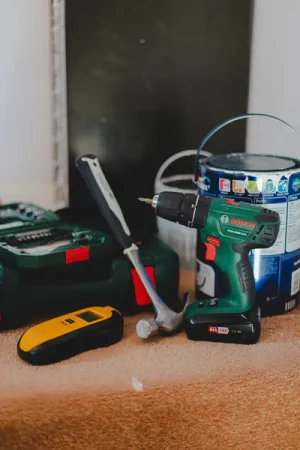 a Bosch PSB 1800 LI-2 cordless drill sits centre stage on a carpeted floor, flanked by a claw hammer, a yellow stud/wire detector, and a large tin of Dulux paint, with a green toolbox case blurred in the background - Practical Maintenance Tips to Keep Your House Running Smoothly and Safely