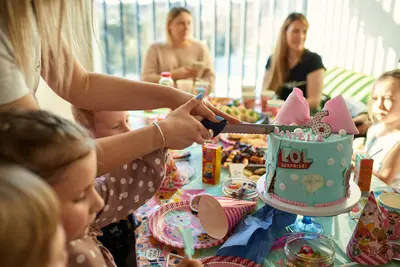 Two hands cut into a pale blue sixth-birthday cake decorated with a pink bow and 'LOL Surprise' design, as children and adults look on at a party table - Unique Hires That Can Make Your Party So Much More Fun