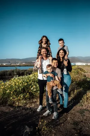A family of six poses together on a wildflower path beside a lake, with two children sitting on the parents' shoulders and a toddler held in arms, under a clear blue sky - Do You Want To Start A Family?