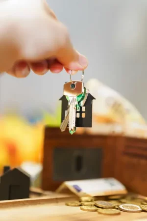 Keys on a house-shaped fob being held up above a wooden table scattered with euro coins, a bank card, and a small black model house, symbolising home ownership or a property purchase. - Unlock Savings Funding for Home Upgrades