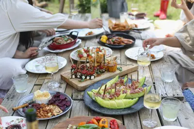 An outdoor wooden table spread with melon-and-prosciutto skewers, grilled vegetable rolls, cheese, nuts and glasses of wine, with people reaching for food in the background - Creative Ways to Enhance Your Outdoor Gatherings