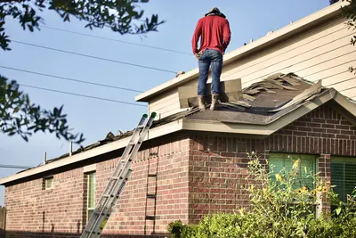 A roofer in a red shirt stands on the ridge of a brick house's roof, surrounded by torn-up shingles, with a ladder leaning against the brickwork below - The Lifespan of Your Roof: Replacement vs. Restoration