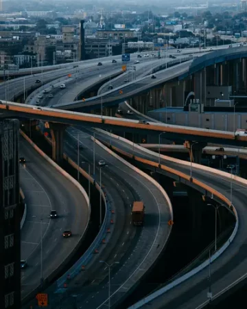 Aerial view of a multi-level urban motorway interchange at dusk, with curving elevated ramps carrying light traffic above a city skyline - How Better Transport Planning Can Support Local Events