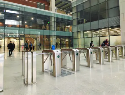 Rows of stainless steel security turnstiles in a glass-walled building atrium, with people passing through in the background - How to Improve Safety in High-Risk and Access-Controlled Environments