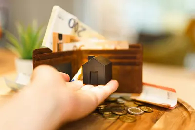 Small model house held in a hand in front of an open wallet with euro banknotes and coins on a table - Selling vs Renting Your Property 4: Things to Consider