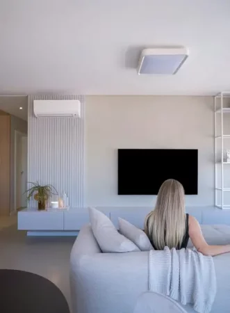 A woman relaxes on a grey sofa in front of a wall-mounted TV, with a wall-mounted air conditioning unit and ceiling light visible above a minimalist floating media console - The Role of Ventilation in Healthy Living Spaces