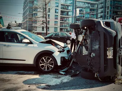 Aftermath of a car accident showing a vehicle flipped on its side beside a badly damaged white Kia, with a tow truck driver assessing the scene - What to Do When an Accident Changes Your Plans
