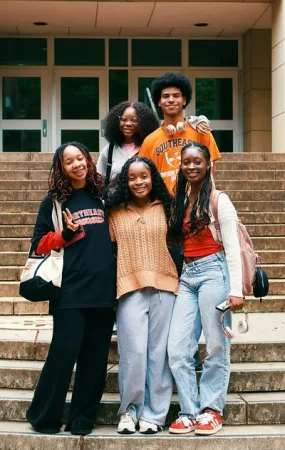 Five smiling students posing together on the stone steps outside a university building entrance - How University Life Shapes Future Community Leaders
