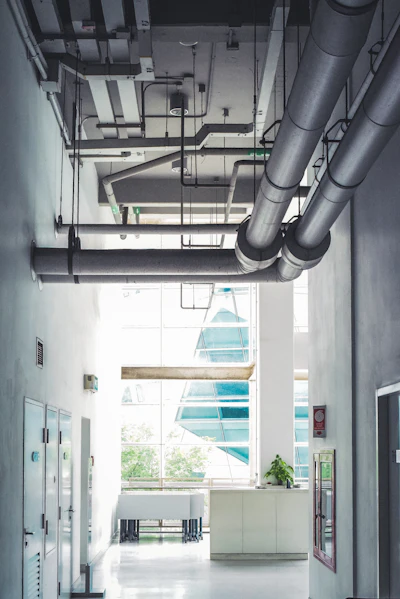 Building Corridor With Overhead Ventilation Ducts - Essential Technical Knowledge for Facilities Management Roles Light-filled building corridor with large insulated ventilation ducts and pipework running along the ceiling above service doors