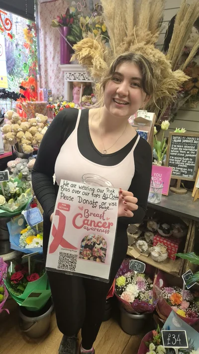 Harriett Evison wearing a pink bra over her uniform and holding the Team Regent Breast Cancer Awareness poster – Bras and Big Hearts: Regent House of Flowers Goes Pink for Breast Cancer Now