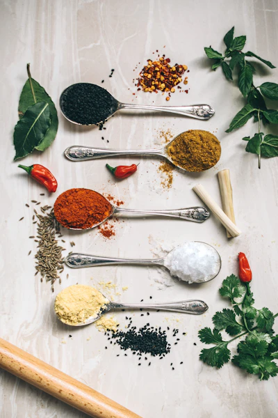 Five vintage spoons filled with black seeds, curry powder, paprika, sea salt and mustard powder beside fresh herbs and red chillies on a marble surface